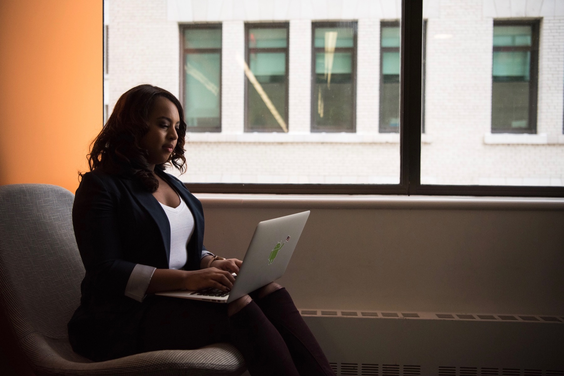 Woman sits in a chair near a window with her computer in her lap.