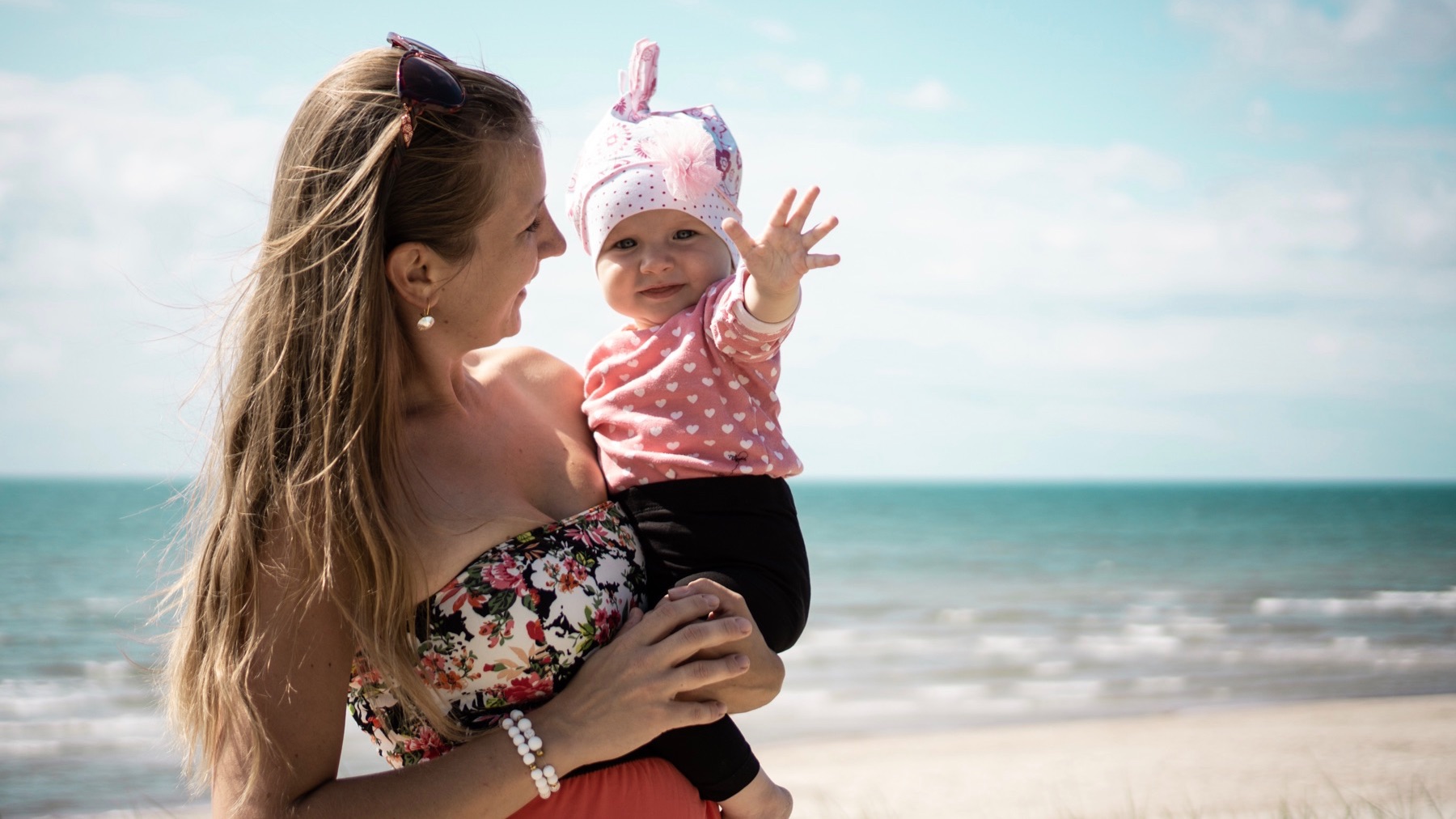 A woman smiles as she holds a baby on a beach near the ocean.