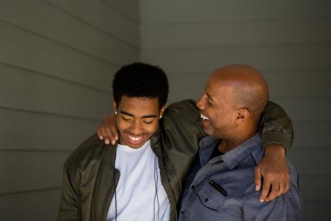 Teenager son and father stand laughing, arms around each other's shoulders