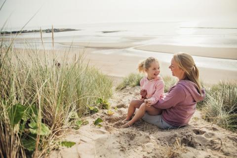 Daughter sits on mother's lap as they relax on a beach.