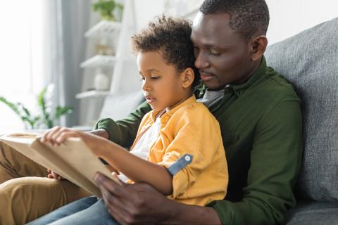 Father holds son in lap on the living room couch while they read their favorite book together.