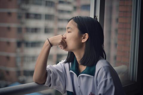 Teenaged girl looks morosely out the window of a high rise apartment.