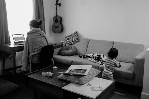 Parent sits at desk, working on laptop, as child plays on the couch behind her.