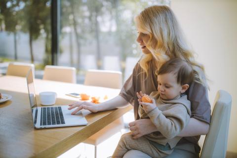 Mom sits down to work on her computer with her child eats a snack while sitting on her lap.