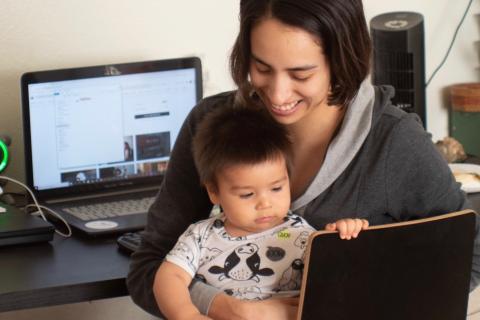 A mother sits at her desk with her child in her lap while she works on her laptop.