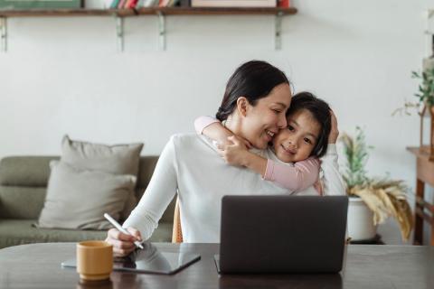 A young child hugs her mother from behind while she works at her computer.
