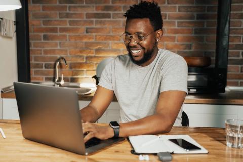 A man smiles as he works on his computer in his kitchen.