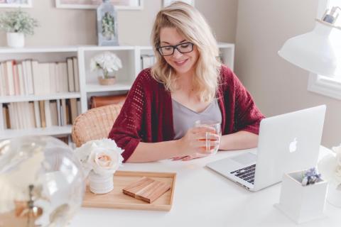 A woman drinks tea as she looks down at her computer and smiles.