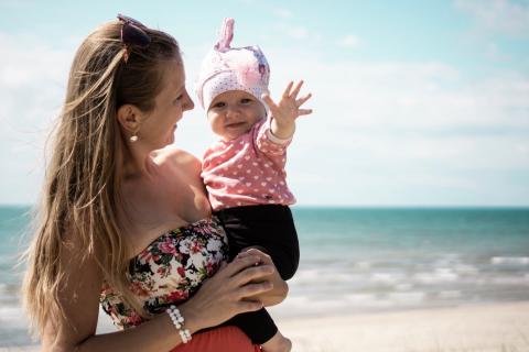 A woman smiles as she holds a baby on a beach near the ocean.