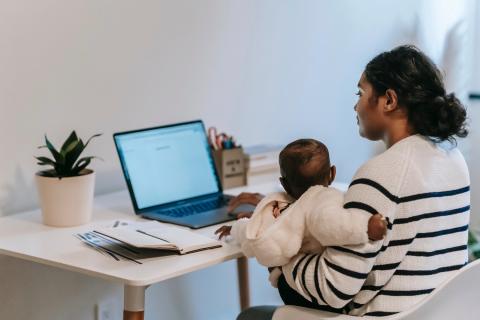 A mother holds an infant on her lap while she uses a laptop at her desk.