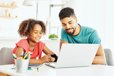 A father and daughter look at a laptop together and smile while sitting at a desk with a cup of pens nearby.