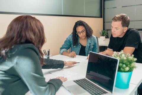 Three adults sit at a table together while one passes paperwork over to the other two to review.