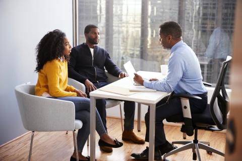 Two adults sit at one end of a table while a third adult reads from a paper on the other end of the table.