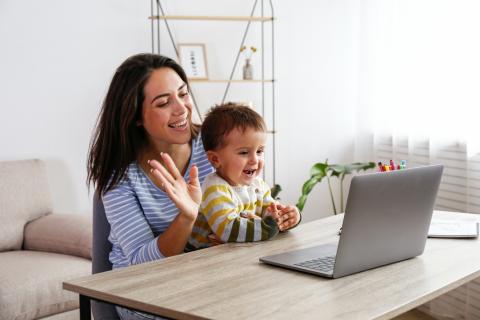 A woman with dark brown hair sits at a table with a young boy in her lap. Both woman and child are smiling and waving at a computer screen.