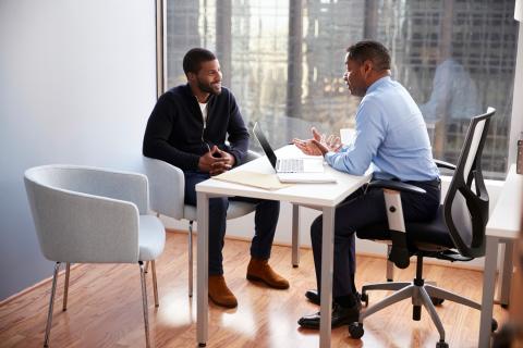 Two men have a discussion while sitting at a table in an office.