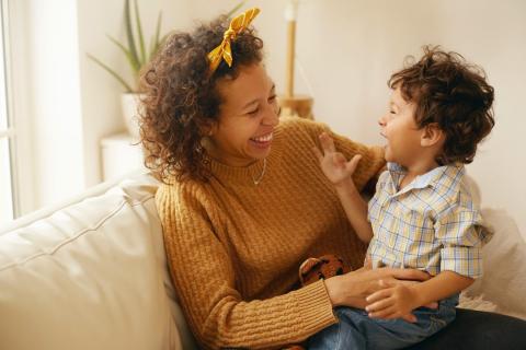 A woman in a mustard yellow sweater holds a young child in her lap while sitting on the couch. They laugh together.