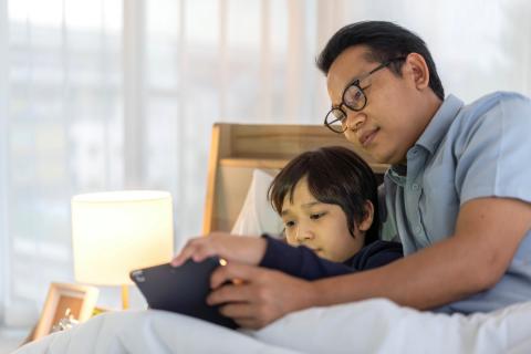 A father and son lay in bed together to read a story on a tablet. 