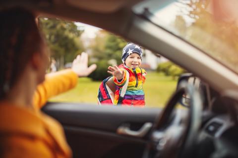 Young boy wearing a backpack waves to his mother outside her car on his way to school.