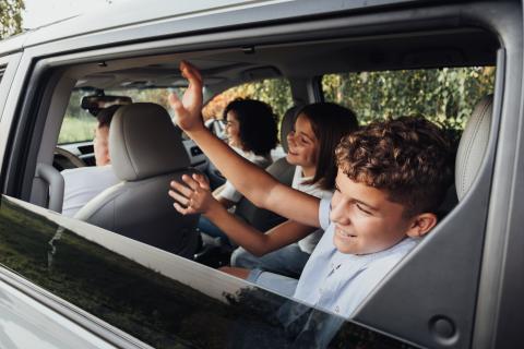 Kids in car waving goodbye to someone outside the vehicle.