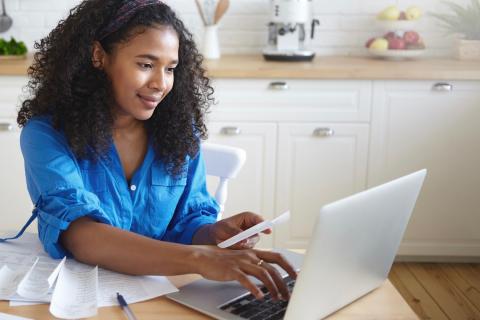 Smiling woman in a blue shirt works on her computer and smartphone at the same time.