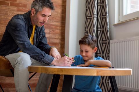 A father and son sit at a small table to draw pictures with colored pencils.