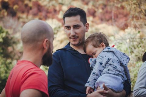 Two dads talking while one holds a sleeping baby. 