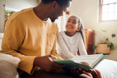 Father and daughter read together while sitting on a bed.