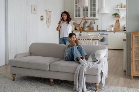 Mom arguing on phone while daughter sits on couch plugging her ears.