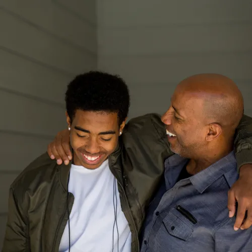 Teenager son and father stand laughing, arms around each other's shoulders