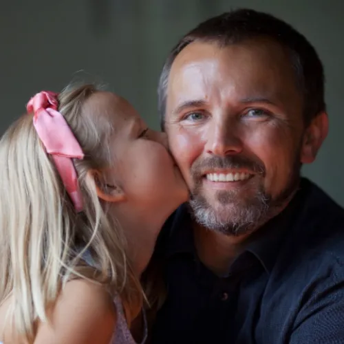 Daughter with a pink bow on her head kisses the cheek of her smiling father