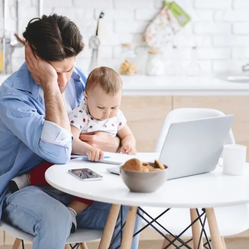 Man sits at his computer desk holding his baby with a disappointed look on his face.
