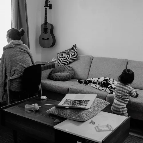 Parent sits at desk, working on laptop, as child plays on the couch behind her.