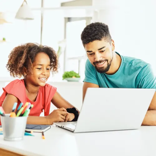 A father and daughter look at a laptop together and smile while sitting at a desk with a cup of pens nearby.