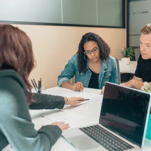 Three adults sit at a table together while one passes paperwork over to the other two to review.