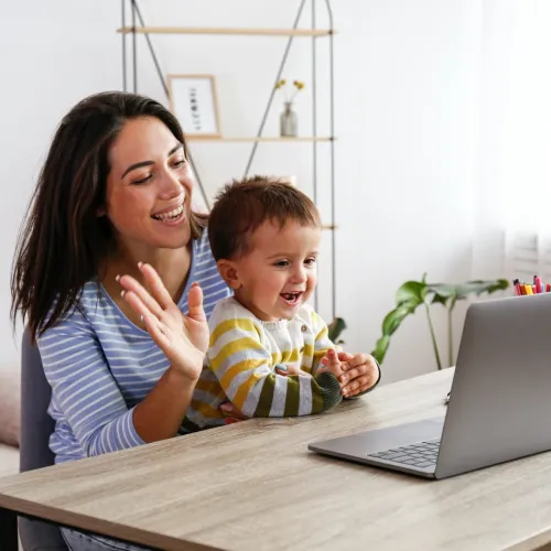 A woman with dark brown hair sits at a table with a young boy in her lap. Both woman and child are smiling and waving at a computer screen.