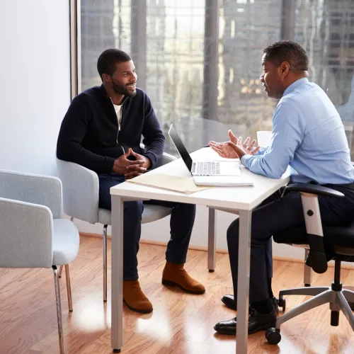 Two men have a discussion while sitting at a table in an office.