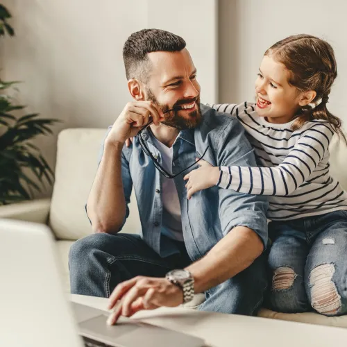 A young girl hugs her father from his side while sitting together on the couch. They look at each other and smile.