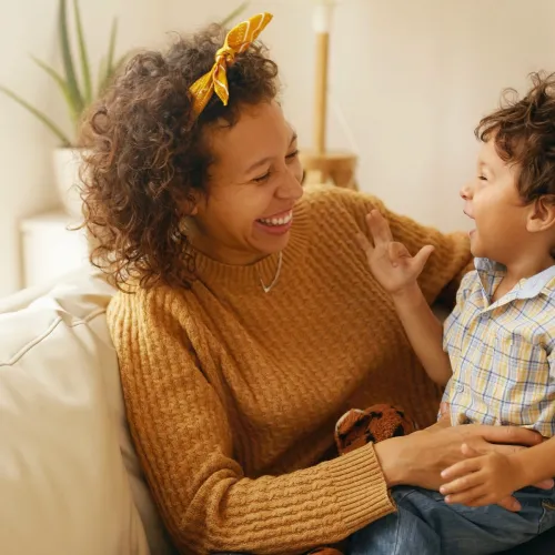 A woman in a mustard yellow sweater holds a young child in her lap while sitting on the couch. They laugh together.