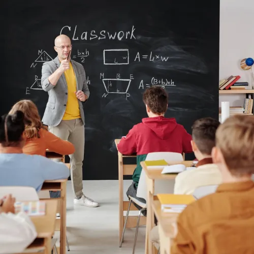 A teacher leading a classroom full of children points to a boy with his hand raised.