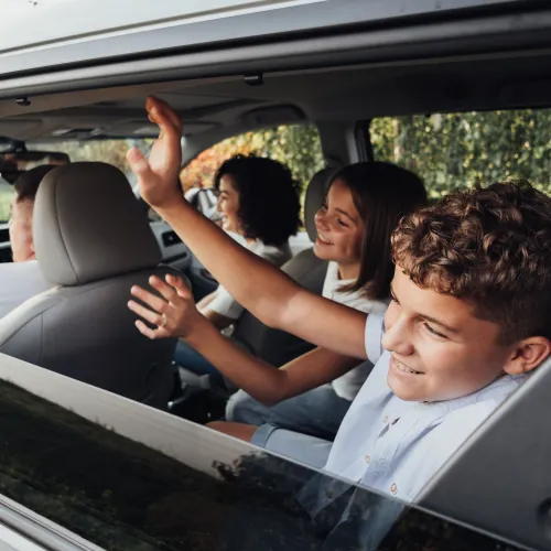 Kids in car waving goodbye to someone outside the vehicle.