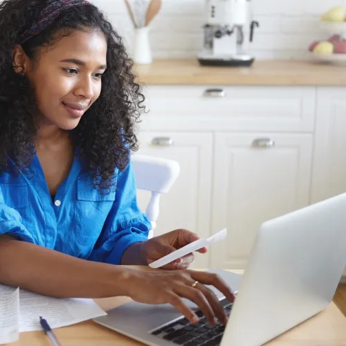 Smiling woman in a blue shirt works on her computer and smartphone at the same time.