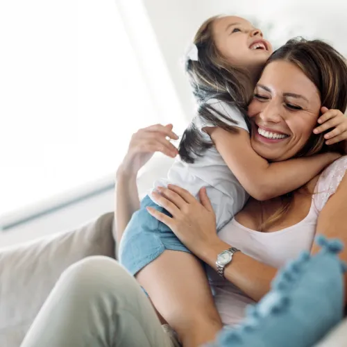A young girl smiles and hugs her mother sitting on a couch.