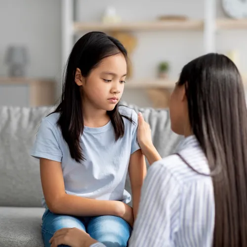 A young girl looks sadly towards the ground as her mother attempts to comfort her.