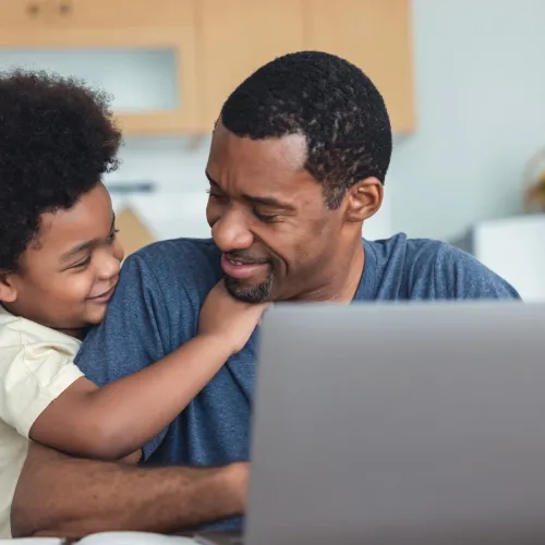 A young boy hugs his father from the size as Dad uses a computer.