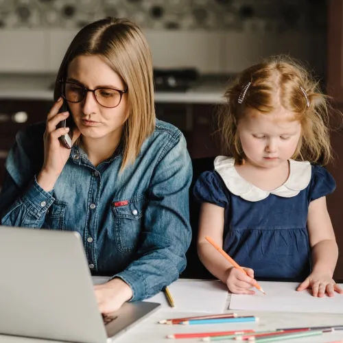 A mother works on a laptop while sitting at a kitchen table with her young daughter drawing on paper beside her.