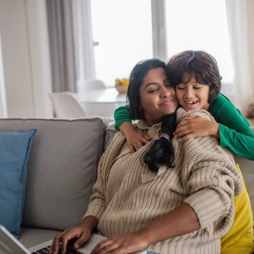 A young boy hugs his mother from behind while she sits on the couch to use her laptop.