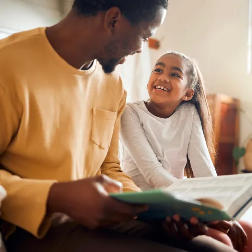 Father and daughter read together while sitting on a bed.