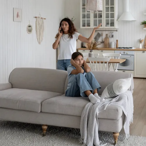 Mom arguing on phone while daughter sits on couch plugging her ears.