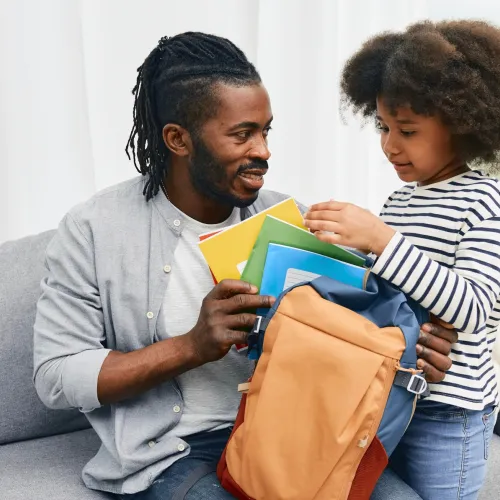 Dad helping young daughter pack her backpack before school.