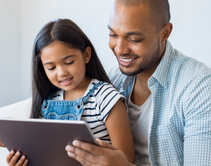 A young girl sits on her father's lap as they look over an electronic tablet together.